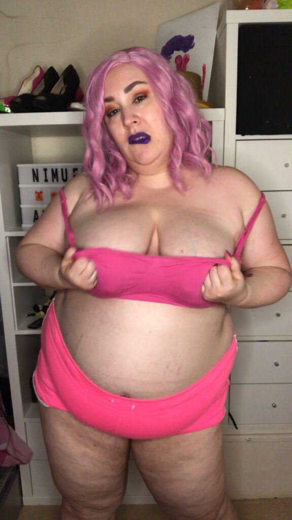 British BBW Nimue Allen wear's a pink wig, pink bra, and pink shorts.  She's pulling down the bra and looking seductively at the camera